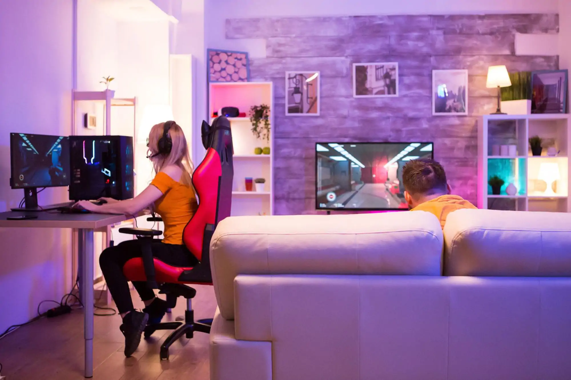 In un salotto illuminato da luci colorate, una ragazza utilizza un computer da gaming con cuffie mentre un ragazzo sul divano guarda e gioca su un grande schermo TV.